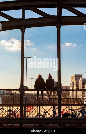 Due giovani donne seduto sulla balaustra del Haccurbrucke ponte sopra i binari ferroviari guardando in lontananza, luce posteriore, Monaco di Baviera, superiore Foto Stock