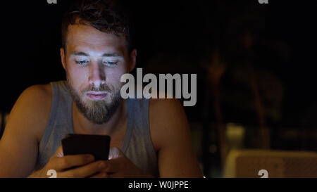 L'uomo utilizza lo smartphone di notte navigando su internet l'aggiornamento di social media. Giovane maschio con la barba utilizzando mobile cell smart phone all'aperto al buio in estate. Foto Stock