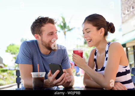 Giovane su cafe guardando smart phone app Foto di bere il caffè in estate. Giovane uomo urbano utilizza lo smartphone sorridendo felice di casual donna asiatica seduti all'aperto. Amici nel tardo 20s Foto Stock