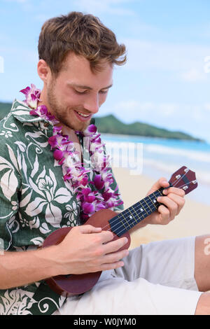 Uomo sulla spiaggia a giocare ukulele strumento su Hawaii. Giovane uomo praticare sulla spiaggia vacanze in abbigliamento hawaiano indossando aloha shirt dress e ghirlanda di fiori. Foto Stock