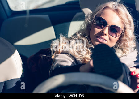 Giovane donna driver in occhiali da sole in ritardo lottando per mettere le scarpe con un piede fino al volante concettuale del disattenzione e Foto Stock