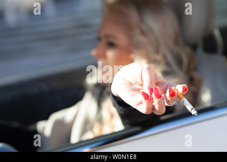 Giovane donna di fumare mentre si guida la sua automobile colpetti ceneri fuori la sua sigaretta attraverso il lato aperto della finestra con la messa a fuoco a mano Foto Stock