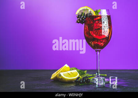 Freddo cocktail rosso con ribes nero, limone, menta e ghiaccio nel bicchiere alto su sfondo viola. In estate i drink e i cocktail alcolico. Cocktail alcolico Foto Stock