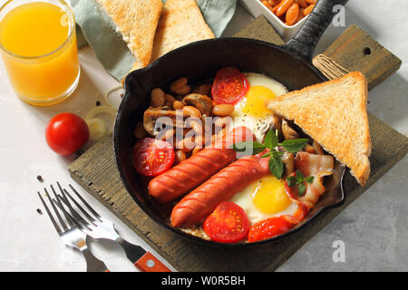 Prima colazione in inglese le uova strapazzate con pancetta, fagioli, funghi, salsicce, pomodori e succo di luce sul tavolo di pietra. Foto Stock