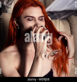 Lonely giovane donna di bere vino in camera d'albergo. redhead donna seduta su un divano con un bicchiere di vino rosso. Foto Stock