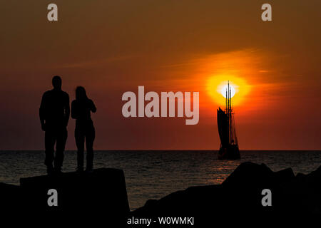 Silhouette di una giovane coppia in piedi sul molo di cemento i blocchi di protezione a Scheveningen rivolto a tall ship passando davanti un vivido momento al tramonto Foto Stock