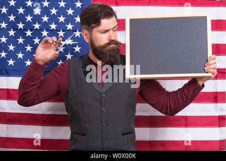 Educazione patriottica. Uomo Barbuto holding forbici e Blackboard nella scuola di educazione. Barbiere corsi di istruzione. Hipster dando lezione sulla bandiera americana sfondo. Istruzione e carriera, copia dello spazio. Foto Stock