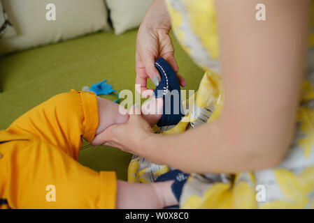 Giovani caucasici madre indossa un vintage abito giallo e aiutando a little baby boy ottenere pronto, con focus sulle mani il montaggio carino scarpe blu Foto Stock