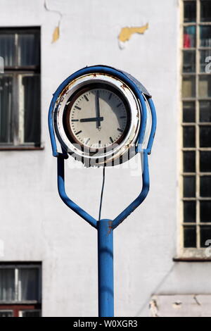 Outdoor orologio analogico con metallo arrugginito telaio in medio di abbandono dei complessi industriali con il vecchio edificio fatiscente in background Foto Stock