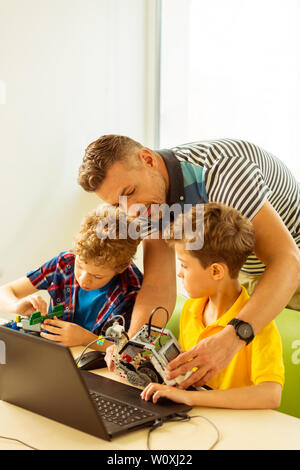 Durante la classe tecnico. Nice helpful insegnante in piedi dietro i suoi allievi mentre li aiutano con il loro progetto Foto Stock