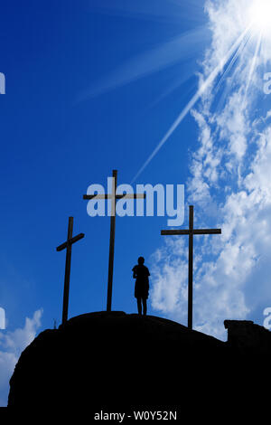 Silhouette di tre croci sulla collina e un ragazzo che prega su un cielo blu con nuvole e raggi solari Foto Stock