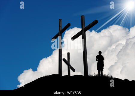 Silhouette di tre croci sulla collina e un ragazzo che prega su un cielo blu con nuvole e raggi solari Foto Stock