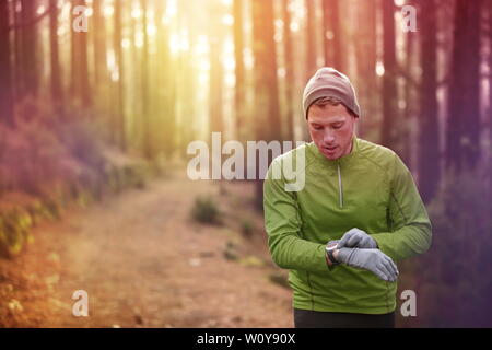 Trail Running runner guardando il monitor per la frequenza cardiaca guardare in esecuzione nella foresta di indossare una giacca calda sportswear, cappello e guanti. Pareggiatore maschio esegue la formazione nel bosco. Foto Stock