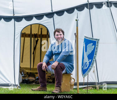 Armadietto medievale rievocativo seduto fuori da una tenda di tela in un evento storico vivente, indossando abiti d'epoca con armi esposte dietro. Foto Stock