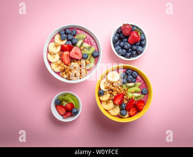 Due Estate acai smoothie ciotole con fragole, banana, mirtilli e kiwi e granola pastello su sfondo rosa. Tazza colazione con frutta e Foto Stock