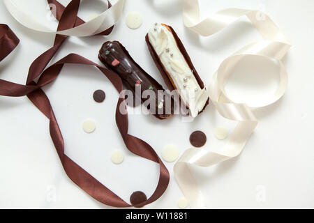 Close-up di un set di due eclairs con diversi ripieni e design isolare su una superficie bianca decorata con gocce di cioccolato, bianco e marrone di seta ri Foto Stock