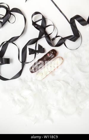 Set di due eclairs con un decor di nero e il cioccolato bianco isolare su una superficie bianca decorata con piume di colore bianco e marrone e nastri, copyspace Foto Stock