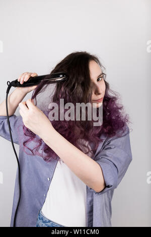 Giovane ragazza si raddrizza i capelli ricci di ferro piatto. La colorazione dei capelli viola. Sfondo grigio Foto Stock