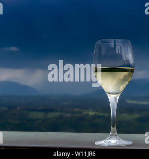 Messa a fuoco selettiva su un bicchiere di vino bianco in un contatore overseing montagne sullo sfondo e si avvicina al sensore pioggia, con abbondanza di spazio copia visto fr Foto Stock