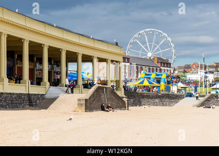La spiaggia di sabbia fine e ruota panoramica Ferris a Barry Island sono illuminate dal sole e si stagliano contro le nuvole scure nel cielo con un inizio di giornata estiva Foto Stock