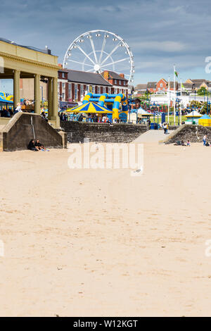 La spiaggia di sabbia fine e ruota panoramica Ferris a Barry Island sono illuminate dal sole e si stagliano contro le nuvole scure nel cielo con un inizio di giornata estiva Foto Stock