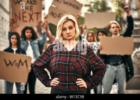 Close up fiducioso femmina giovane militante con un gruppo di persone sul retro per protestare e tenendo la segnaletica. Gruppo protestando all'esterno. Le donne di marzo Foto Stock