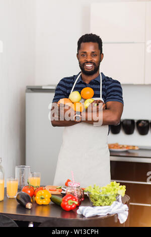 Attraente sorridente giovane barbuto dark scuoiati uomo che indossa il grembiule tenendo fresche e mature arance organico, mele e succo di pompelmo, rendendo frullato di frutta, godeteveli Foto Stock