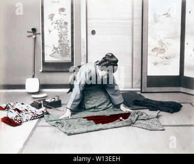 [ 1890 Giappone - donna giapponese il ripiegamento di un kimono ] - una donna in kimono e acconciatura tradizionale è il ripiegamento di un kimono. Altri kimono si posa sul pavimento. Vi è un tubo di kiseru, casella di tabacco e una shamisen dietro di lei. Xix secolo albume vintage fotografia. Foto Stock