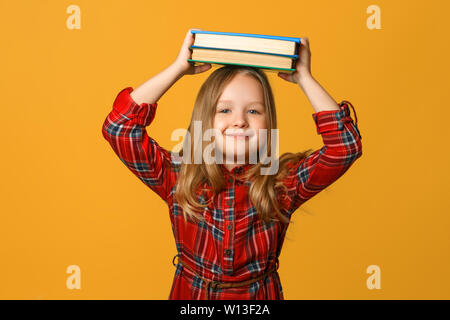 Ritratto di una bambina schoolgirl su sfondo giallo. Il bambino tiene libri sulla sua testa. Il concetto di istruzione e la scuola. Foto Stock