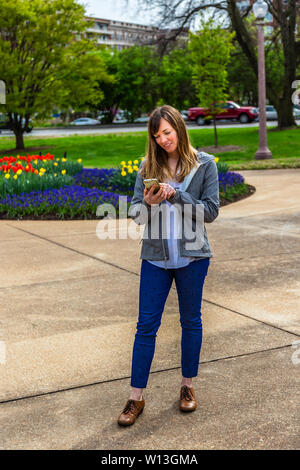 Giovane donna in piedi che guarda verso il basso flowersat il suo telefono in un parco. Foto Stock