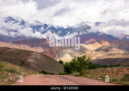 La strada verso le montagne colorate e vette innevate sullo sfondo di un cielo nuvoloso. Viaggiare in Kirghizistan Foto Stock