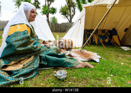 Medievale storia vivente rievocazione. Maiden, donna, nel XII secolo il verde ed il giallo del costume seduti all'aperto sull'erba dalla tenda medievale. Vista laterale. Foto Stock