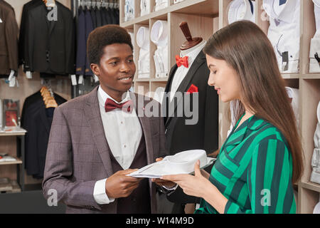 Ragazza, consulente shop scegliendo una camicia bianca per client di eleganti boutique. Venditore camicia di contenimento in mani e mostra all'uomo. Ampia scelta di shir bianco Foto Stock