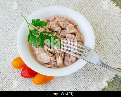 Conserve di tonno filetto in porcellana bianca bowl, forcella, prezzemolo e alcuni pomodori ciliegia su un beige tabella tabella igienico. Frutti di mare, mangiare sano. Vista dall'alto. Foto Stock