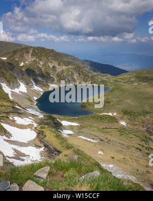 Iconico vista dal picco dei laghi sulla montagna Rila presso il lago di rene con rocce in primo piano, altopiani soleggiati ed erba verde e drammatiche le nuvole in un cielo Foto Stock