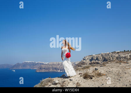 Bella ragazza in un abito bianco sorge su una scogliera e guarda al mare, la Grecia, l'isola di Santorini. Foto Stock