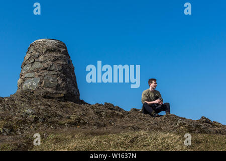 Giovane età 20 Siede alla Scottish highland mountain top a fianco di pietra cairn, l'uomo guardando in lontananza con cielo blu senza nuvole Foto Stock