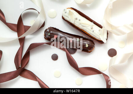 Close-up di un set di due eclairs con diversi ripieni e design isolare su una superficie bianca decorata con gocce di cioccolato, bianco e marrone di seta ri Foto Stock