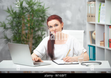 Fiducioso medico donna seduta al tavolo nel suo ufficio e lavora al computer. Il concetto di assistenza sanitaria. Foto Stock