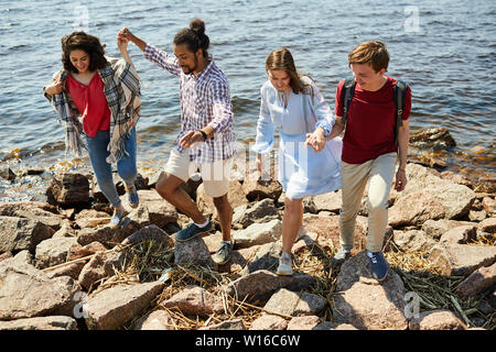 A piena lunghezza ritratto di due giovani coppie camminando sulla spiaggia rocciosa e dal mare, spazio di copia Foto Stock