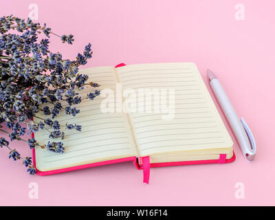 Fragrante bouquet di lavanda a secco con piccoli fiori viola su un aperto blocco di carta in un coperchio di colore rosa e bianco con penna a sfera su una rosa pastello backgro Foto Stock