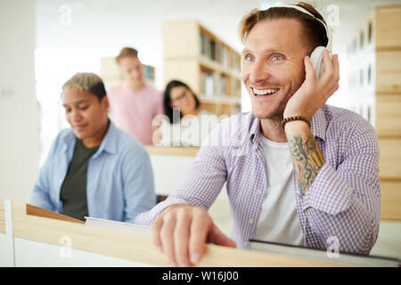 Eccitati positivo bel ragazzo con tatuaggio sul braccio in piedi al bancone di accoglienza e ascolto di musica in cuffia in libreria Foto Stock