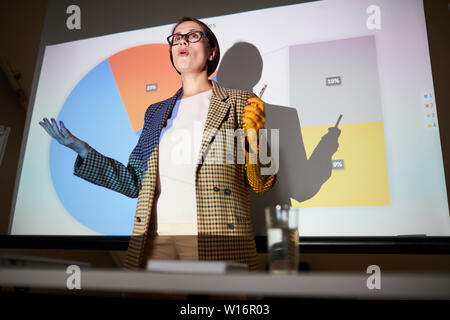 Al di sotto di vista del fiducioso donna matura in camicia a scacchi gesti delle mani mentre spiegare i dati su schermo di proiezione in classe di formazione Foto Stock