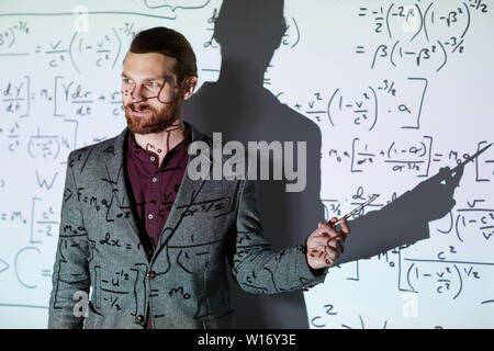 Contenuto fiducioso giovane barbuto insegnante di matematica in camicia in piedi contro lo schermo di proiezione e puntando alla formula spiegando i calcoli Foto Stock