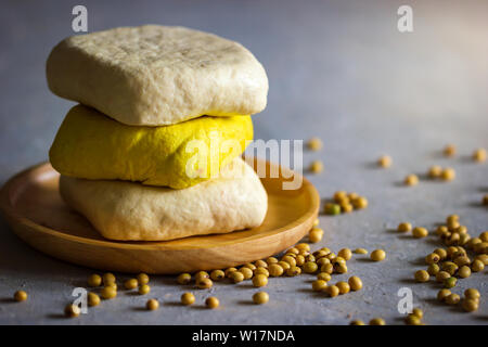 Il tofu impilamento sul piatto di legno con la luce del sole di mattina e fagioli di soia semi sparsi sul tavolo. Cibo sano concetto. Foto Stock