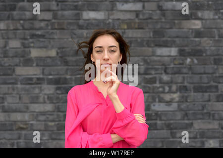 Premurosa donna in piedi a fissare la fotocamera con la mano al suo mento e una espressione contemplativa nella parte anteriore del grigio di un muro di mattoni Foto Stock