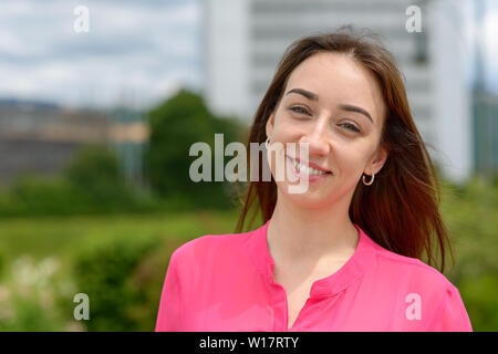 Felice attraente giovane donna con un bel caloroso sorriso amichevole in piedi fuori nel sole grinning presso la fotocamera Foto Stock