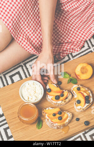 Ragazza del picnic a mangiare la frutta bruschetta con crostini di baguette, fresco morbido di ricotta, succose pesche, mirtilli e miele. Concetto di estate, salute Foto Stock