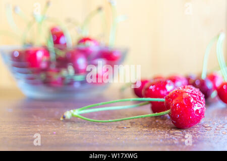 Red ciliegie mature in una ciotola di vetro mantenuta su un tavolo di legno in cucina. L'acqua spruzzata su di ciliegie fresche mantenuto sul tavolo di legno. Messa a fuoco selettiva di ch Foto Stock