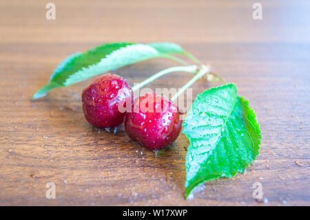 Appena prelevato red ciliegie mature con foglie mantenuta su un tavolo di legno con acqua spruzzata su di essi. Foto Stock